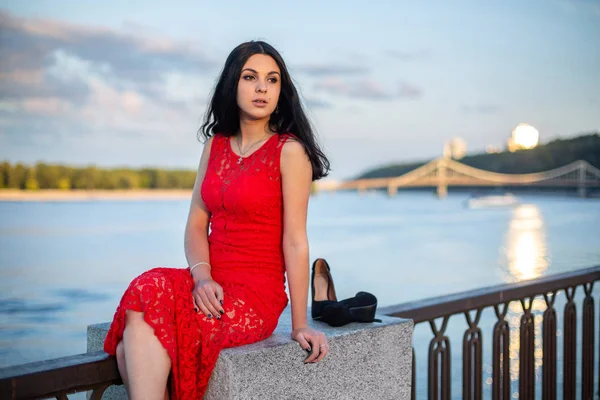 Una chica vestida de rojo está sentada en la cerca del terraplén del río. . — Foto de Stock