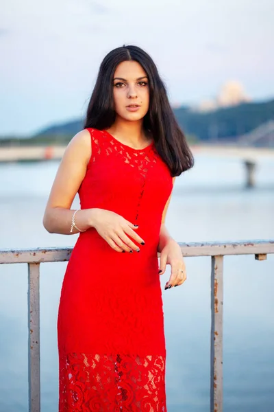 Hermosa joven con un vestido rojo posando en un puente cerca de una vieja cerca . —  Fotos de Stock