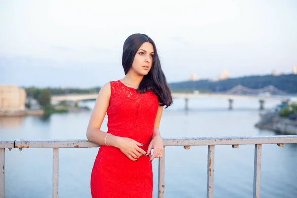 Hermosa joven con un vestido rojo posando en un puente cerca de una vieja cerca . —  Fotos de Stock