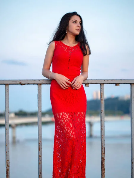 Menina bonita em um vestido vermelho posando em uma ponte perto de uma cerca velha . — Fotografia de Stock