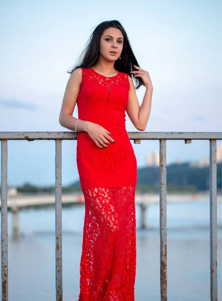 Menina bonita em um vestido vermelho posando em uma ponte perto de uma cerca velha . — Fotografia de Stock