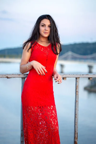 Menina bonita em um vestido vermelho posando em uma ponte perto de uma cerca velha . — Fotografia de Stock