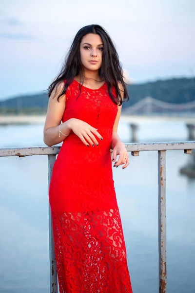 Menina bonita em um vestido vermelho posando em uma ponte perto de uma cerca velha . — Fotografia de Stock