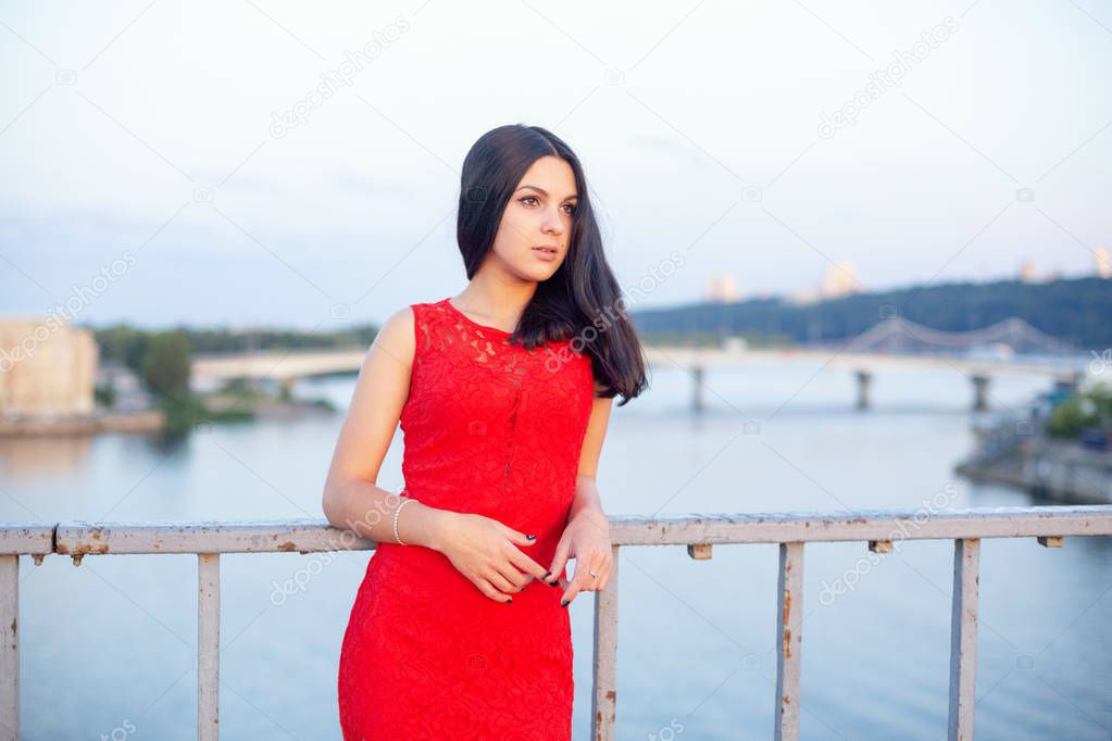 Beautiful young girl in a red dress posing on a bridge near an old fence.