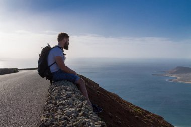 Bir adam yaz giysileri bir taş çit oturan ve okyanusa yakın bir ada bakar. Mirador del Rio, Lanzarote, İspanya.