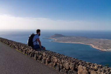 Bir adam yaz giysileri bir taş çit oturan ve okyanusa yakın bir ada bakar. Mirador del Rio, Lanzarote, İspanya.