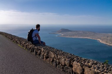 Bir adam yaz giysileri bir taş çit oturan ve okyanusa yakın bir ada bakar. Mirador del Rio, Lanzarote, İspanya.