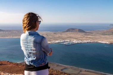 Bir kadın bir uçurumun kenarında duruyor ve okyanusa yakın bir ada bakar. Rüzgar saç katlar. Mirador del Rio, Lanzarote, İspanya.