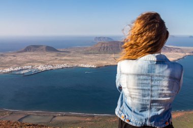 Bir kadın bir uçurumun kenarında duruyor ve okyanusa yakın bir ada bakar. Rüzgar saç katlar. Mirador del Rio, Lanzarote, İspanya.