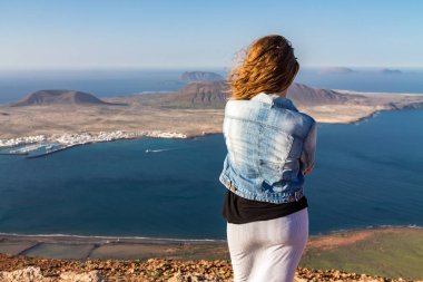 Bir kadın bir uçurumun kenarında duruyor ve okyanusa yakın bir ada bakar. Rüzgar saç katlar. Mirador del Rio, Lanzarote, İspanya.