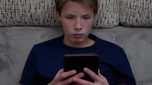 En pojke i blå t-shirt sitter på soffan och läser en e-bok. — Stockvideo