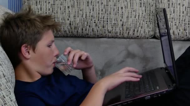 Der Junge benutzt einen Laptop und trinkt Wasser. — Stockvideo