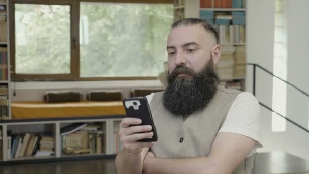 Молодий чоловік з бородою щось читає на своєму смартфоні і має вау-реакцію — стокове відео