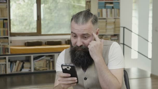 Reação de um jovem com barba tentando entender e pensar no que ele vê em sua tela de telefone inteligente — Vídeo de Stock