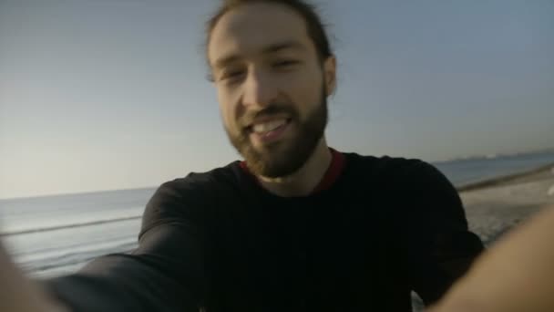 Крупный план портрета счастливого человека с длинными волосами и бородой, держащего камеру на пляже, пытающегося сделать видео селфи своим отпуском — стоковое видео