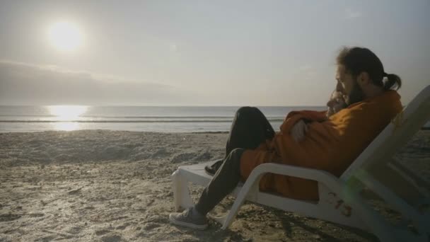 Hermosa pareja joven cubierta con una manta abrazándose y acostada en un sillón en la playa disfrutando de la puesta de sol — Vídeos de Stock