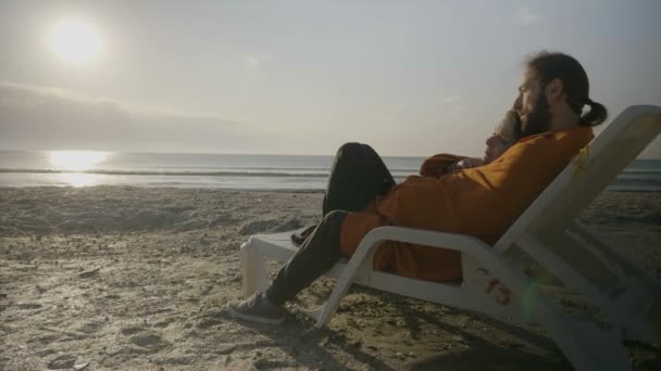 Ласковая молодая пара, лежащая на шезлонге на пляже и смотрящая на закат под одеялом — стоковое видео