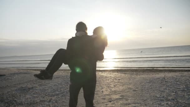 Joven pareja feliz en el amor arremolinándose en la playa hombre sosteniendo mujer en sus brazos mientras el sol se pone al atardecer en cámara lenta — Vídeos de Stock