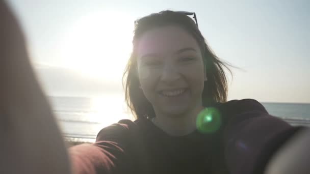 Lächelnde schöne Frau mit Sonnenbrille, die ein Selfie mit dem Handy am Strand macht, während sie sich dreht und das Meer in Zeitlupe genießt — Stockvideo