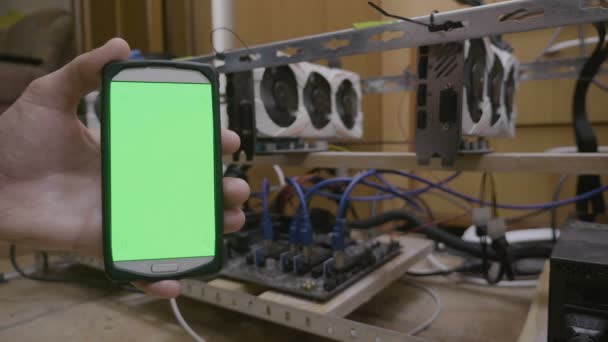 コードを入力する画面に触れる開発者トレーダー男主催 Cryptocurrency 採掘リグの横にある緑色の画面を持つスマート フォン表示します — ストック動画