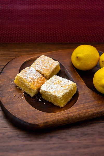 Home baked lemon squares, lemon bars, with lemons over wooden blurred background in portrait orientation