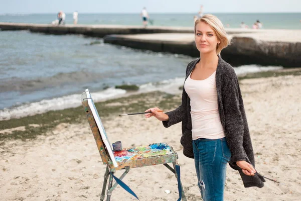 A young woman artist paints a landscape on the beach. — Stock Photo, Image