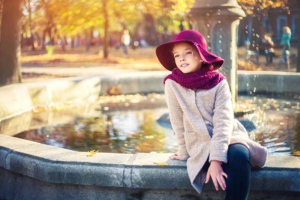 Flicka i klassisk kappa och mössa i höst parken nära fontänen. Höstsäsongen, mode, barndom. — Stockfoto