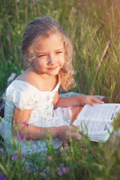 Little girl with a book in her hands on a meadow in a summer day. — Stock Photo, Image