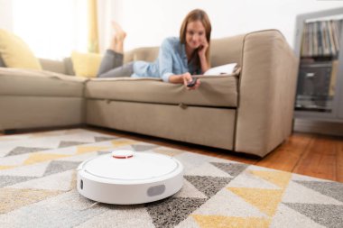 Kadın kanepede dinlenirken robot süpürge odayı temizliyor. Uzaktan kumandayla vakumu kontrol eden kadın.