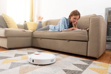 Kadın kanepede dinlenirken robot süpürge odayı temizliyor. Uzaktan kumandayla vakumu kontrol eden kadın.