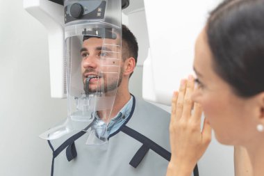 Patient standing in x-ray machine at dental clinic clipart