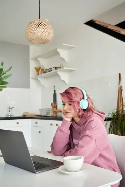 Hipster teen girl pink hair using laptop sit at kitchen table in chair vertical.