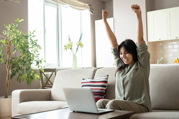 Excited indian woman celebrating online win looking at laptop at home.