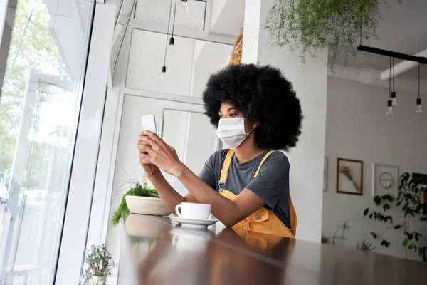 African lady wearing face mask using phone sit at table in modern cafe.