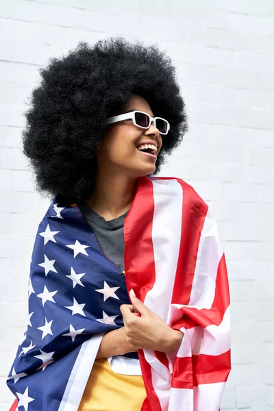Happy young African woman wrapped in USA flag standing against white wall.