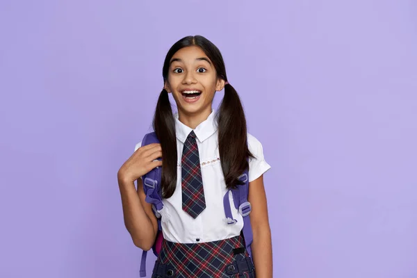 Excited amazed indian school girl holds backpack isolated on violet background.