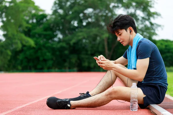 Young Asian athletic man sitting and listening to music from mobile phone to relax during rest