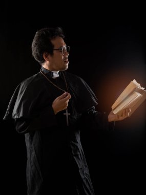 Gözlüklü genç rahip eski İncil 'i tutuyor ve siyah arka planda dua ediyor.
