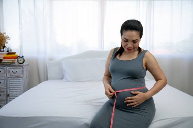 Koca göbekli, gri elbiseli, hamile, Asyalı obez kadın. Yatakta otururken bantla karnını şişiriyor.