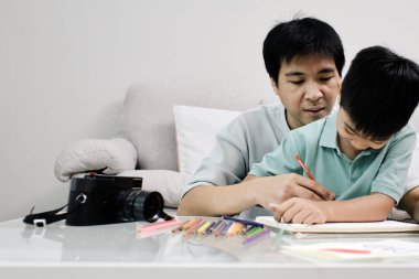 Baba, küçük oğluna oturma odasındaki renkli kalemlerle resim yapmasını öğretiyor.