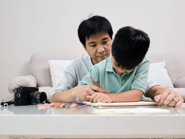 Babam kanepede oturmuş, oturma odasındaki renkli kalemlerle çizdiği resimlerle oğlana rehberlik ediyor.