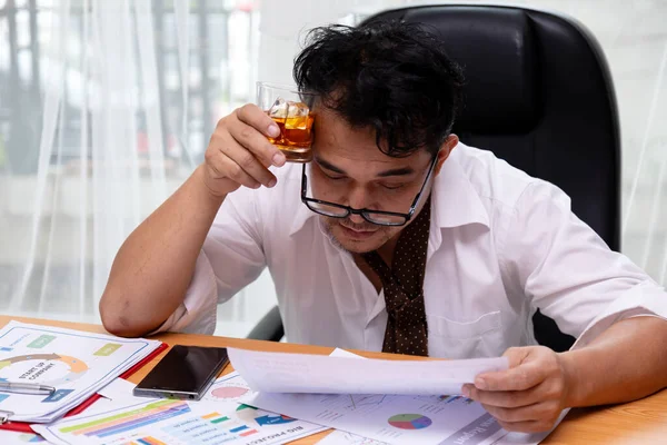 Gevşemiş kravatlı işadamı viski içerken mali tabloları ve raporları analiz ederek stresi ve işyerindeki tükenmeyi sembolize ediyor.