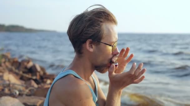 穿着衬衣和太阳镜的年轻人在海边的口琴上演奏 — 图库视频影像