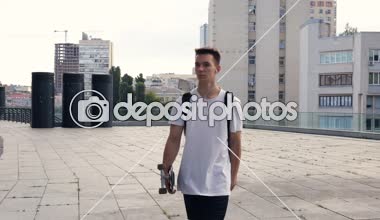 Kaykay holding, yürüyüş ve müzik genç adam. Sabit cam vurdu.