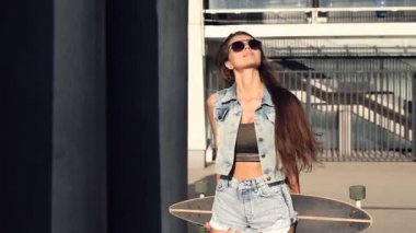 Poz ve sokakta ayakta longboard olan kadın. Kameraya bakarak.