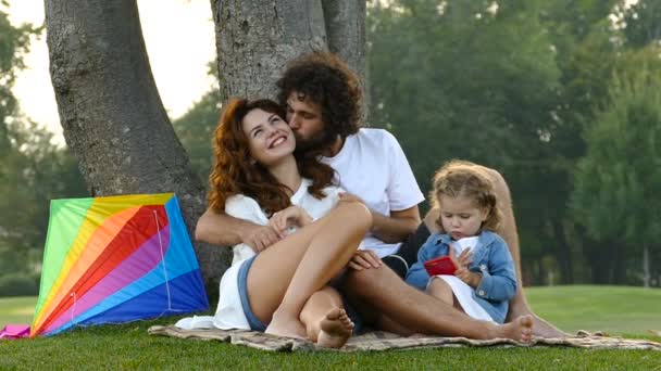 父の母と小さな娘が公園の芝生の上に座っています ハグの夫妻 娘持株スマート フォンと再生します スローモーション — ストック動画