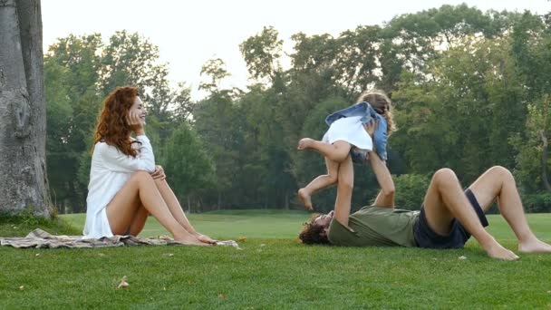 在城市公园的树附近的家庭 妻子看着丈夫 在草地上和小女儿玩耍 Slowmotion — 图库视频影像