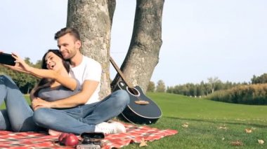 Genç çift ekose ağacın yanında oturan. Selfies üstünde smartphone alarak ve having fun.