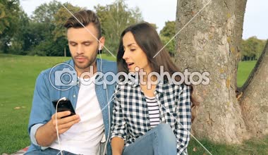 Genç çift açık havada. Ekose ve Smartphone müzik parçasını seçme ağacın yanında oturan.