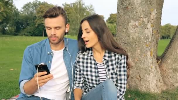 年轻夫妇在户外 坐在格子上的树上 选择智能手机上的音乐播放列表 — 图库视频影像